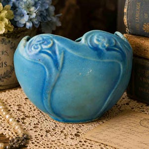 May include: A bright turquoise ceramic vase with a rounded, heart-like shape and embossed floral relief patterns along the upper rim. The vessel features a smooth, matte-textured finish and a wide, open top, showcasing a vibrant blue color.