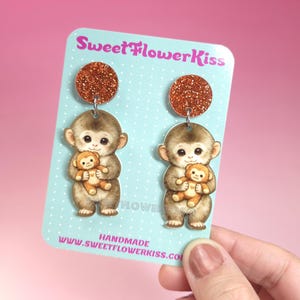 May include: Dangle earrings with a brown glitter top and a monkey holding a teddy bear. The earrings are on a light blue card with the text "SweetFlowerKiss" and "HANDMADE".