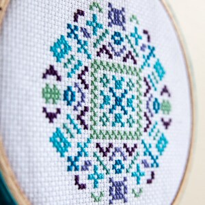 May include: A white embroidery hoop with a teal blue trim, featuring a cross-stitch design of a geometric pattern in shades of blue, green, and purple.