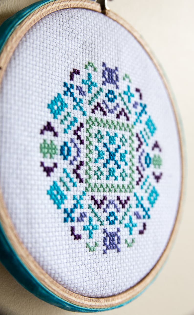 May include: A white embroidery hoop with a teal blue trim, featuring a cross-stitch design of a geometric pattern in shades of blue, green, and purple.