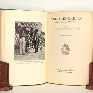 May include: An open book titled "The Rain-Makers" by Mary Roberts Coolidge. The book features a black and white photograph of a Native American ceremony. The book is bound in blue and is propped up by two red bookends.