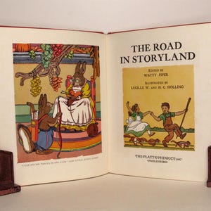 May include: An open book titled "The Road in Storyland" with illustrations of a rabbit with a bow and arrow and two children walking in a field. The book is edited by Watty Piper and illustrated by Lucille W. and H. C. Holling. The book is published by The Platt & Munk Co. Inc.