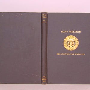 May include: An open book with a dark gray cover and spine. The title "MANY CHILDREN" is in gold lettering, along with the author's name, "MRS SCHUYLER VAN RENSSELAER." A gold emblem is centered on the cover.
