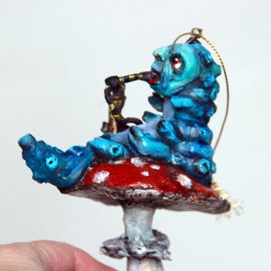 May include: A decorative ornament depicting a blue caterpillar with a telescope, sitting on a red and white mushroom. The caterpillar has a detailed, textured design. The ornament is suspended by a gold string, suitable for hanging.