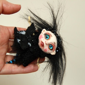 Puede incluir: Una muñeca hecha a mano con una cara pálida, ojos azules y labios rojos. La muñeca tiene cabello negro y lleva un atuendo negro con una falda de malla. La muñeca está decorada con cuentas azules y detalles blancos.
