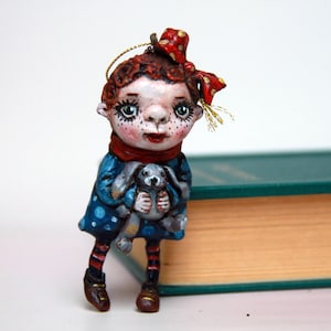 Könnte beinhalten: Eine handgefertigte Puppe mit rotem Haar, einer roten Schleife und einem blauen Kleid mit weißen Punkten. Die Puppe hält ein graues Häschen und trägt einen roten Schal. Die Puppe steht neben einem grünen Buch.