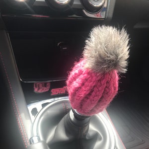 May include: A pink knitted car gear shift knob with a gray faux fur pom pom on top.