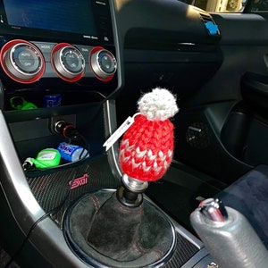 K&ouml;nnte beinhalten: Ein roter und wei&szlig;er gestrickter Automatik-Schalthebelknauf mit einem wei&szlig;en Bommel oben. Der Knauf ist an einem schwarzen Schalthebel in einem Autoinnenraum befestigt.