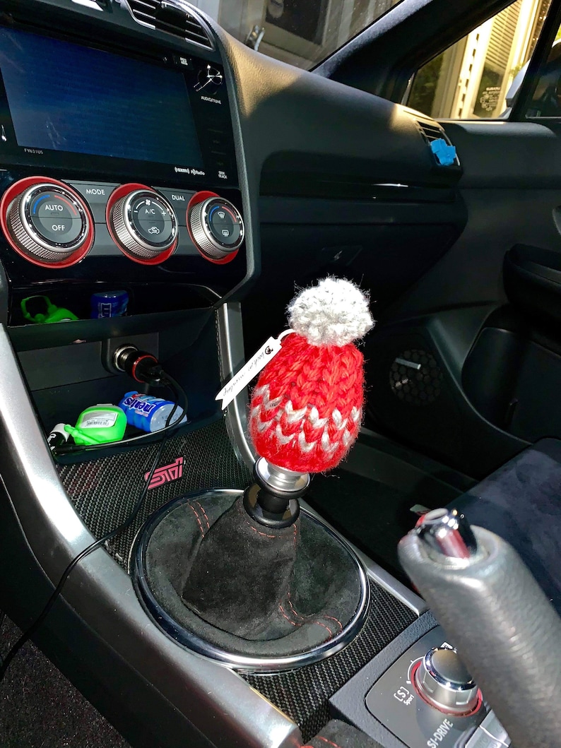K&ouml;nnte beinhalten: Ein roter und wei&szlig;er gestrickter Automatik-Schalthebelknauf mit einem wei&szlig;en Bommel oben. Der Knauf ist an einem schwarzen Schalthebel in einem Autoinnenraum befestigt.