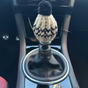 K&ouml;nnte beinhalten: Ein schwarzer und beiger gestrickter Schaltknauf f&uuml;r ein Auto mit einem schwarzen Bommel oben. Der Knauf befindet sich in einem Auto mit schwarzem Lederausstattung und roten N&auml;hten.