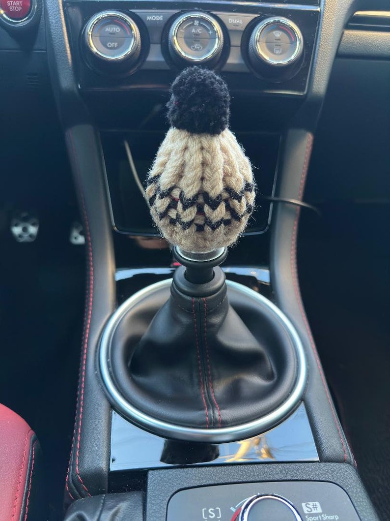 K&ouml;nnte beinhalten: Ein schwarzer und beiger gestrickter Schaltknauf f&uuml;r ein Auto mit einem schwarzen Bommel oben. Der Knauf befindet sich in einem Auto mit schwarzem Lederausstattung und roten N&auml;hten.