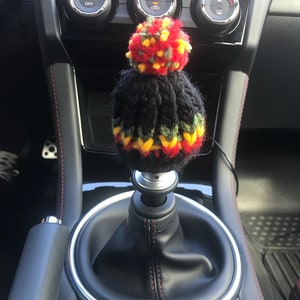 K&ouml;nnte beinhalten: Ein schwarzer gestrickter Schaltknauf mit einem roten, gelben und gr&uuml;nen Bommel oben. Der Knauf befindet sich in einem Auto mit schwarzem Lederausstattung.