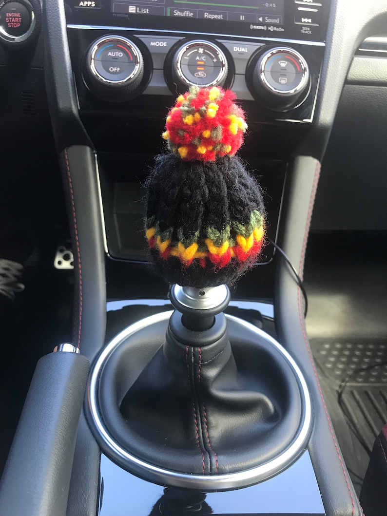K&ouml;nnte beinhalten: Ein schwarzer gestrickter Schaltknauf mit einem roten, gelben und gr&uuml;nen Bommel oben. Der Knauf befindet sich in einem Auto mit schwarzem Lederausstattung.