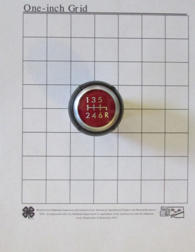 K&ouml;nnte beinhalten: Ein schwarzer Schaltknauf mit roter Oberseite. Die Oberseite hat wei&szlig;e Zahlen 1, 3, 5, 2, 4, 6 und den Buchstaben R.