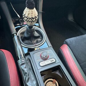 K&ouml;nnte beinhalten: Eine schwarz-wei&szlig;e Strickm&uuml;tze mit einem schwarzen Bommel sitzt auf dem Schalthebel eines Autos. Der Innenraum des Autos ist schwarz mit roten Ledersitzen. Das Auto hat einen schwarzen Lederschalthebelbalg und einen schwarzen Lederhandbremsbalg. Das Auto hat eine schwarze Kunststoff-Mittelkonsole mit einem silbernen Knopf und einem silbernen Knopf. Das Auto hat einen schwarzen Kunststoff-Cupholder mit einem braunen geh&auml;kelten Untersetzer.