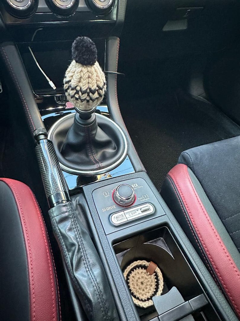 K&ouml;nnte beinhalten: Eine schwarz-wei&szlig;e Strickm&uuml;tze mit einem schwarzen Bommel sitzt auf dem Schalthebel eines Autos. Der Innenraum des Autos ist schwarz mit roten Ledersitzen. Das Auto hat einen schwarzen Lederschalthebelbalg und einen schwarzen Lederhandbremsbalg. Das Auto hat eine schwarze Kunststoff-Mittelkonsole mit einem silbernen Knopf und einem silbernen Knopf. Das Auto hat einen schwarzen Kunststoff-Cupholder mit einem braunen geh&auml;kelten Untersetzer.