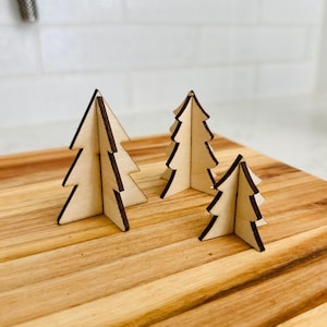 May include: Three wooden Christmas tree decorations of varying sizes. The trees are made of light-colored wood with dark brown edges, and are arranged on a wooden cutting board. The background is a white tiled wall.
