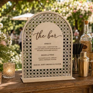 May include: An arched wooden bar menu sign featuring a light-colored cane webbing border. The sign displays the text "the bar" in script, followed by lists for spirits, beers, wine, and soft drinks. It stands on a rectangular wooden base.