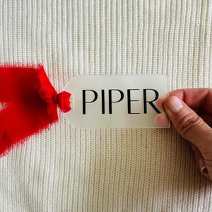 May include: A white gift tag with the name "PIPER" printed in black lettering. The tag is held by a hand against a white knitted background. A red ribbon is partially visible in the bottom left corner of the image.