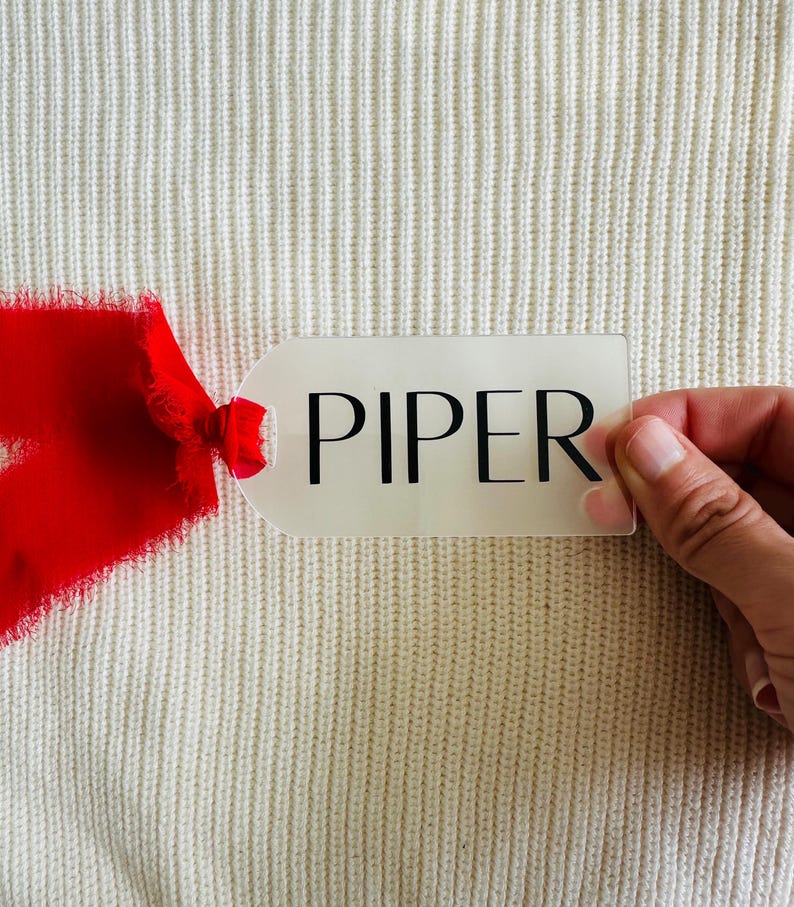 May include: A white gift tag with the name "PIPER" printed in black lettering. The tag is held by a hand against a white knitted background. A red ribbon is partially visible in the bottom left corner of the image.