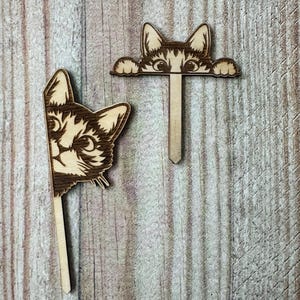 May include: Two wooden cat head ornaments with a stick attached to the back. The cat heads are peeking over the top of a wooden surface. The cat heads are brown and have black eyes and whiskers.