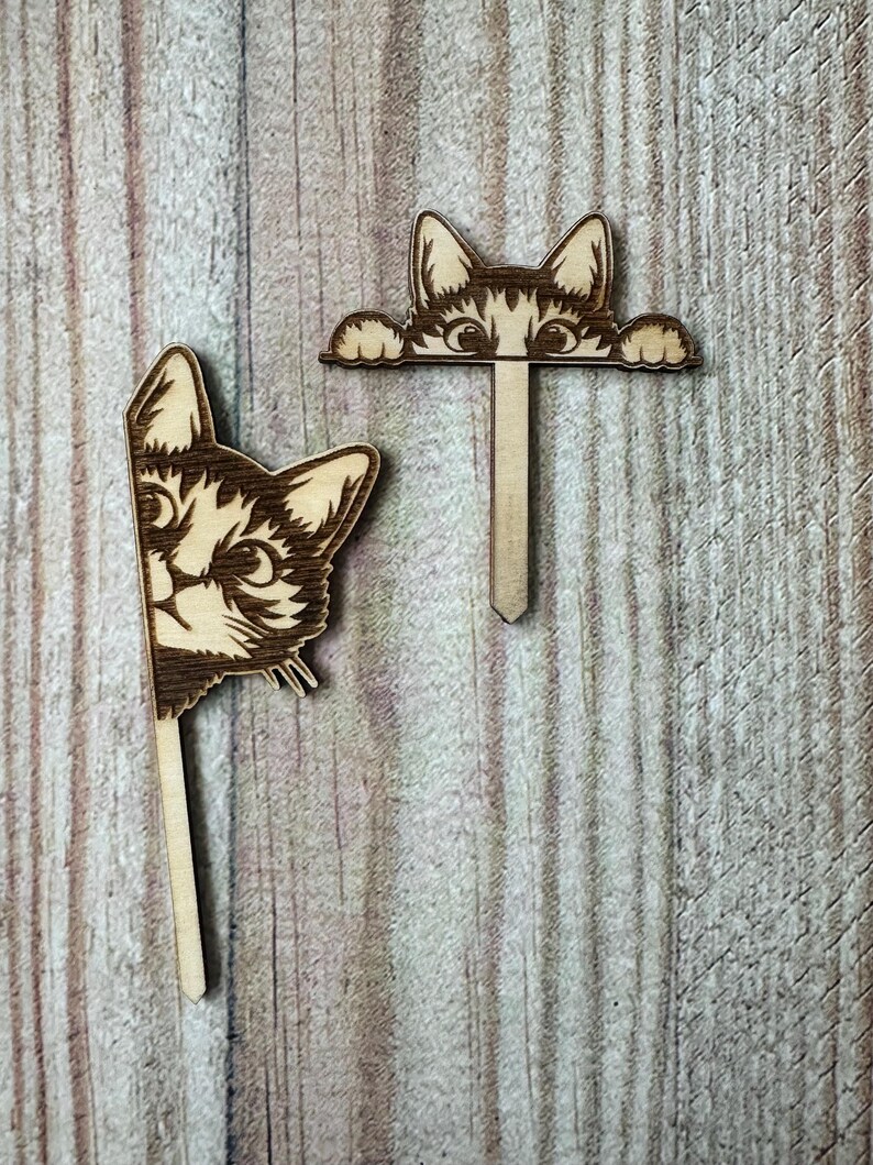 May include: Two wooden cat head ornaments with a stick attached to the back. The cat heads are peeking over the top of a wooden surface. The cat heads are brown and have black eyes and whiskers.