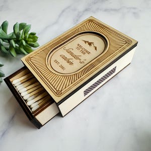 Pu&ograve; includere: Scatola di fiammiferi in legno con cassetto scorrevole, incisa con la scritta "Welcome to the Sander's cabin" e un grafico di montagna. La scatola &egrave; aperta, rivelando fiammiferi con punte bianche. Il design include linee decorative e l'anno "EST. 2001".