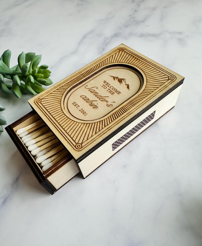 Pu&ograve; includere: Scatola di fiammiferi in legno con cassetto scorrevole, incisa con la scritta "Welcome to the Sander's cabin" e un grafico di montagna. La scatola &egrave; aperta, rivelando fiammiferi con punte bianche. Il design include linee decorative e l'anno "EST. 2001".