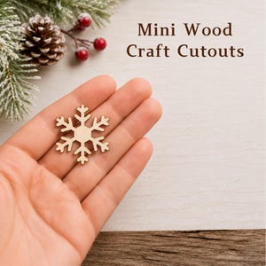 May include: A light-colored wooden snowflake cutout with a six-pointed design. The text "Mini Wood Craft Cutouts" is printed in dark brown letters. The snowflake is held in a palm, with a pinecone and red berries visible in the upper corner.