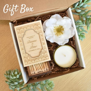 Pu&ograve; includere: Confezione regalo contenente una scatola di fiammiferi in legno con i nomi "Dianne & Peter" e la data "10 ottobre 2025", un fiore bianco e una piccola candela in una scatola dorata. La scatola &egrave; riempita di carta marrone.
