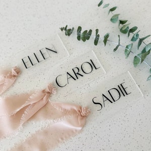 May include: Three clear acrylic place cards with the names "Ellen", "Carol", and "Sadie" written in black font. The place cards are decorated with light pink frayed ribbon.