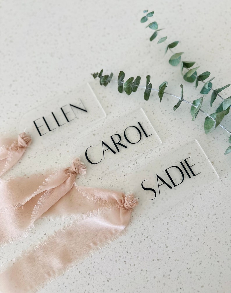 May include: Three clear acrylic place cards with the names "Ellen", "Carol", and "Sadie" written in black font. The place cards are decorated with light pink frayed ribbon.