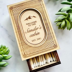 Pu&ograve; includere: Scatola di fiammiferi in legno con un design decorativo e la scritta "Welcome to the Sander's cabin EST. 2001". La scatola &egrave; aperta, rivelando fiammiferi con punte bianche. La scatola &egrave; circondata da piante grasse verdi.