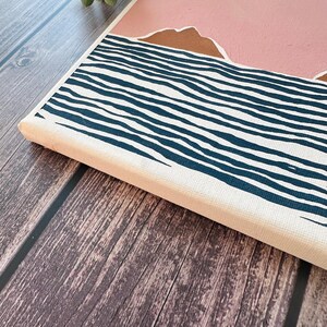 May include: A canvas painting depicting a pink sky with brown mountains above a blue and white striped sea.