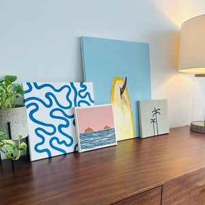 May include: Four framed art prints with a beach theme. The prints feature a blue and white abstract design, a pink sunset with two islands, a yellow surfboard, and a grey print with two palm trees.