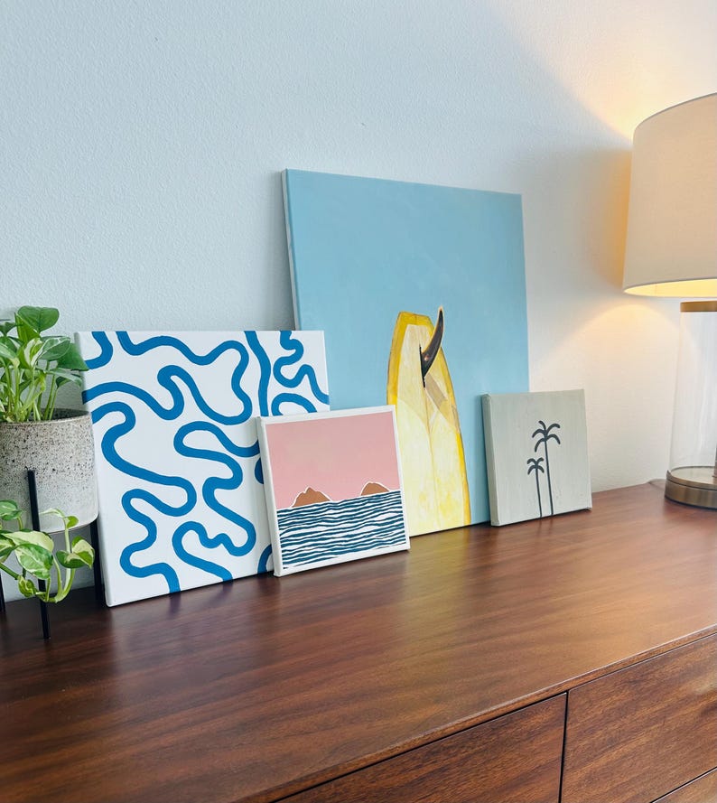 May include: Four framed art prints with a beach theme. The prints feature a blue and white abstract design, a pink sunset with two islands, a yellow surfboard, and a grey print with two palm trees.