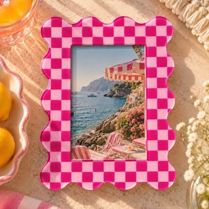 May include: A rectangular picture frame with a wavy, scalloped edge featuring a bright pink and light pink checkerboard pattern. The frame holds a photograph of a coastal scene with a striped umbrella, lounge chairs, and rocky cliffs by the sea.