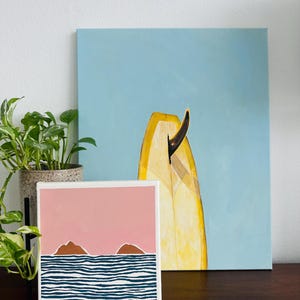 May include: A small painting depicting a pink sky with two brown islands and blue and white striped waves. The painting is on a white canvas and is positioned on a wooden table in front of a larger painting of a yellow surfboard against a blue sky.