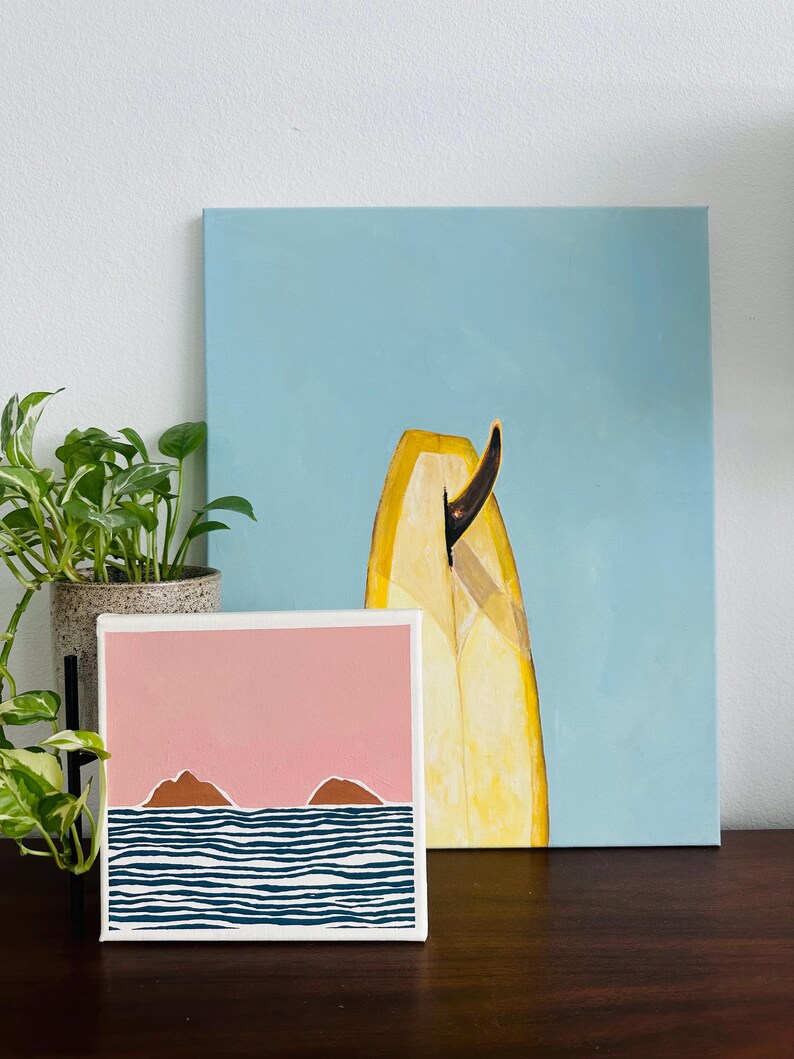 May include: A small painting depicting a pink sky with two brown islands and blue and white striped waves. The painting is on a white canvas and is positioned on a wooden table in front of a larger painting of a yellow surfboard against a blue sky.