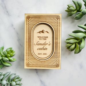 Pu&ograve; includere: Un cartello in legno con la scritta "Welcome to the Sander's cabin EST. 2001" in una cornice ovale. Il cartello presenta una grafica di montagna e un bordo decorativo. Il cartello &egrave; in legno chiaro.
