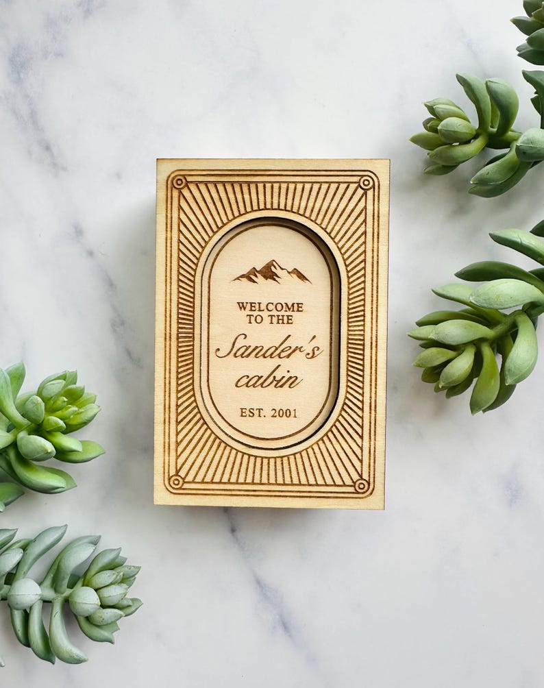 Pu&ograve; includere: Un cartello in legno con la scritta "Welcome to the Sander's cabin EST. 2001" in una cornice ovale. Il cartello presenta una grafica di montagna e un bordo decorativo. Il cartello &egrave; in legno chiaro.