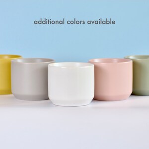 May include: Five ceramic planters in different colors: yellow, gray, white, pink, and green.  The planters are arranged in a row on a white surface.  "additional colors available" is written in black text on a light blue background.