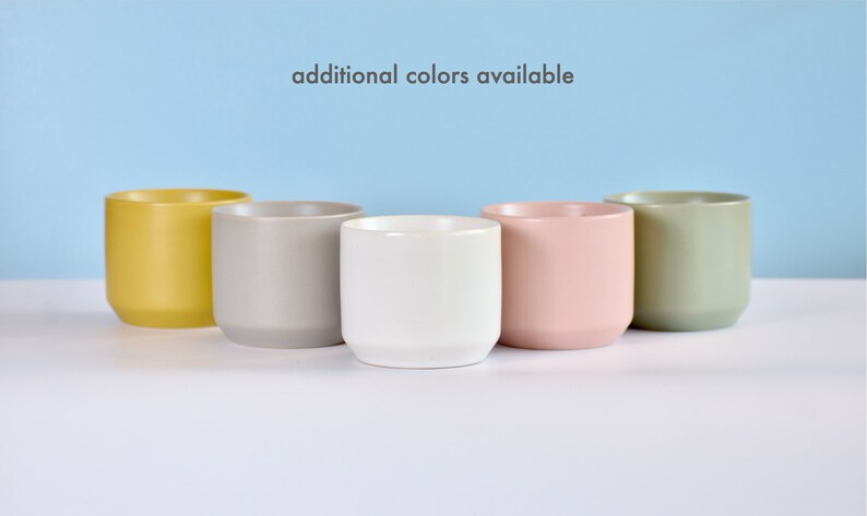 May include: Five ceramic planters in different colors: yellow, gray, white, pink, and green.  The planters are arranged in a row on a white surface.  "additional colors available" is written in black text on a light blue background.