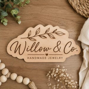 Può includere: Una targa in legno chiaro con bordo smerlato che presenta un ramo di foglie inciso nella parte superiore. Il testo recita Willow & Co. in corsivo, seguito da un piccolo cuore e dalle parole HANDMADE JEWELRY in lettere maiuscole.