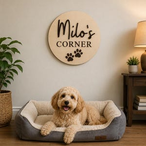 Può includere: Una targa da parete rotonda in legno chiaro con il testo nero Milo's CORNER e due impronte di zampe scure. Sotto la targa, un cane dal pelo chiaro e riccio riposa in una cuccia rettangolare di colore grigio e crema.