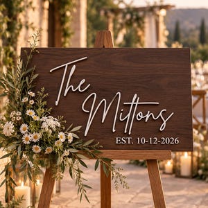 May include: A rectangular dark brown wooden sign resting on a wooden easel. The sign features white 3D lettering that reads The Mittons and the text EST. 10-12-2026. A floral arrangement of white daisies and green foliage is attached to the left side.