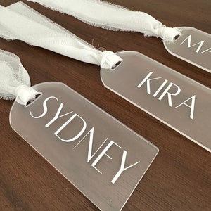 May include: Three clear acrylic tags with white ribbon tied around them. The tags have the names "Sydney", "Kira", and "Max" written on them in white.