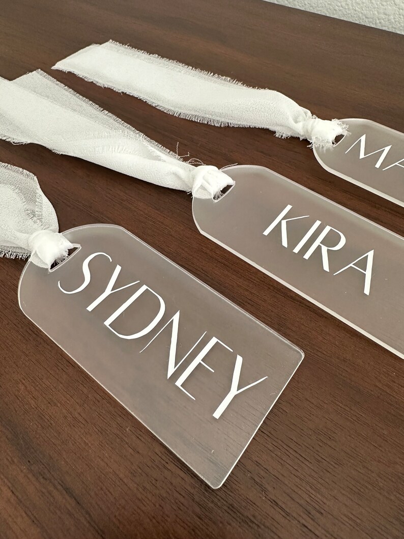 May include: Three clear acrylic tags with white ribbon tied around them. The tags have the names "Sydney", "Kira", and "Max" written on them in white.