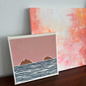 May include: A small canvas painting with a pink background and two brown hills in front of a blue and white striped ocean.