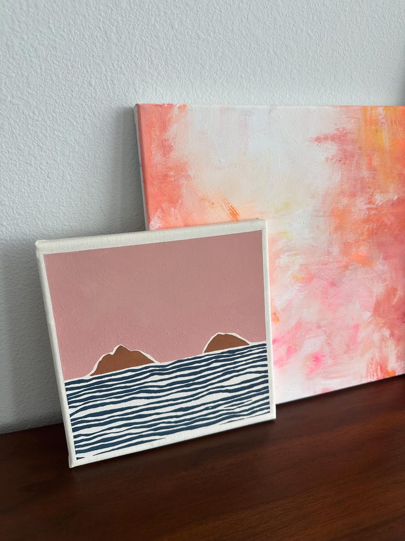 May include: A small canvas painting with a pink background and two brown hills in front of a blue and white striped ocean.